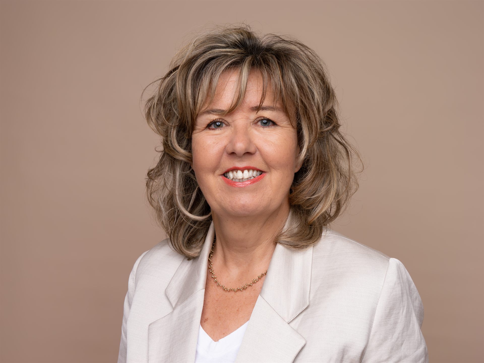 Zakelijke headshot van een glimlachende vrouw in een lichte blazer tegen een beige achtergrond.